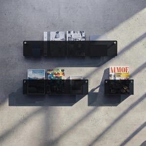 Könnte beinhalten: Drei schwarze, wandmontierte Acryl-Schweberegale mit silbernen Beschlägen. Das obere Regal enthält zwei Bücher. Das untere linke Regal enthält zwei Bücher und einen kleinen Rahmen. Das untere rechte Regal enthält ein Magazin namens AIMOE SHNEE SHNE.