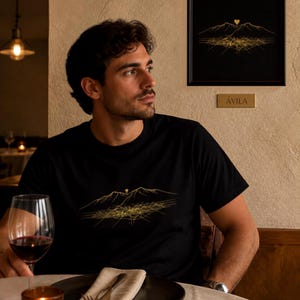 Puede incluir: Una camiseta negra con un gráfico de líneas doradas que representa una cordillera y una cuadrícula urbana, con un pequeño icono de corazón sobre los picos. El mismo diseño aparece en un marco negro con una placa que dice ÁVILA.