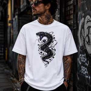 Puede incluir: Una camiseta blanca de manga corta con un estampado gráfico central de una serpiente negra enroscada con ojos y detalles en violeta brillante, rodeada de esferas oscuras flotantes y líneas abstractas en espiral en el frente.