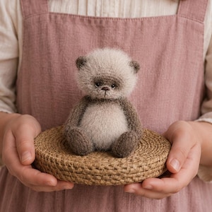 Può includere: Un piccolo orsacchiotto artigianale e soffice con testa color crema chiaro e arti e orecchie marrone scuro. L'orso ha un corpo rotondo e peluche, occhi scuri e un naso cucito, seduto su una base rotonda, intrecciata e di colore marrone chiaro.