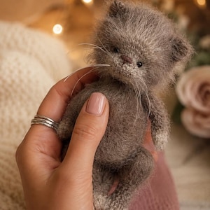 Könnte beinhalten: Eine kleine, flauschige, graue Kätzchenfigur aus Garn mit einer texturierten, weichen Optik. Die Miniaturkatze hat dunkle Augen, eine kleine braune Nase und lange, dünne weiße Schnurrhaare, die aus der Schnauze ragen und feine Handarbeit zeigen.