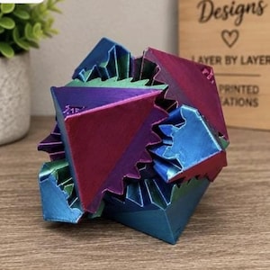 May include: A multi-faceted geometric puzzle object featuring a metallic, iridescent finish that shifts between shades of deep blue, vibrant purple, and emerald green. The structure consists of interlocking triangular and jagged components.