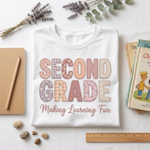 Può includere: Una t-shirt bianca con la scritta "SECOND GRADE" in grandi lettere stampatello decorate con motivi floreali, a quadri e a cuori in tonalità rosa, viola e beige. Sotto, il testo "Making Learning Fun" è scritto in carattere corsivo.