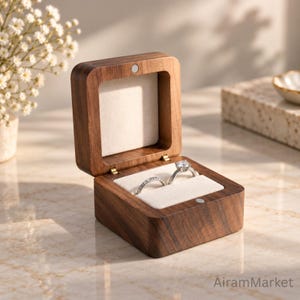 May include: A square dark brown wooden ring box with a hinged lid and a magnetic closure. The interior is lined with soft cream-colored fabric, featuring two slots holding a silver-toned band and a silver-toned ring with a clear stone setting.