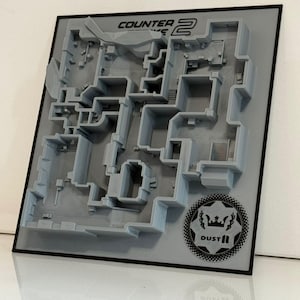 May include: A gray 3D-printed relief map of the Counter-Strike 2 Dust II game level on a square board. The top features the text Counter-Strike 2, and the bottom right corner displays a circular emblem with a crown, laurel, and the text DUST II.