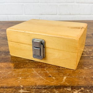 May include: A rectangular light-colored wooden box with a smooth finish and a silver-toned metal latch centered on the front. The box features a hinged lid and a simple, clean construction, showcasing the natural grain and texture of the wood.