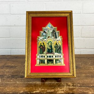 May include: A rectangular gold-toned frame containing a multi-tiered, arched religious illustration set against a vibrant red fabric background. The artwork features detailed figures in various colors, including blue, green, and gold, arranged in a structured, architectural composition with multiple levels and decorative elements.