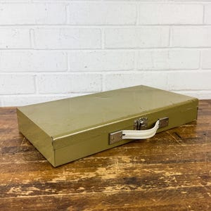 May include: A rectangular olive green metal storage case featuring a white plastic handle and a silver-toned metal latch closure. The exterior surface of the box shows minor signs of wear and surface scratches across the top lid.