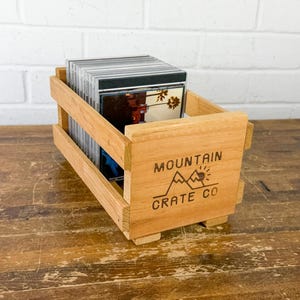 May include: A light brown wooden crate with open slats, designed for storage. The front panel features a black printed logo with the text MOUNTAIN CRATE CO and a mountain graphic. The crate is filled with a row of compact disc cases.