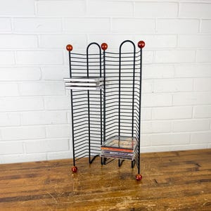 May include: A black wire CD storage rack with a folding design and rounded top arches. The rack features small, spherical, reddish-brown wooden finials at the top and bottom of each vertical support post, holding several compact disc cases.