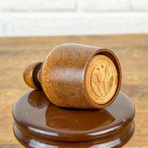 May include: A dark brown wooden stamp featuring a carved tulip design on its circular face. The stamp has a rounded handle and a cylindrical body, showcasing a visible wood grain texture throughout the dark stained surface of the tool.