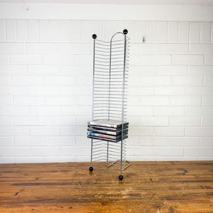 May include: A tall, slender media storage rack constructed from thin, silver-toned metal wire. The unit features a vertical grid design with four small black spherical caps at the top and bottom corners. A small stack of five DVDs rests on the rack.