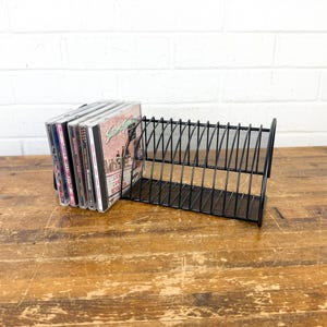 May include: A black metal wire rack designed for organizing compact discs. The rack features a series of vertical dividers to hold multiple cases upright. A small collection of music CDs is stored on the left side of the open storage unit.