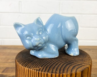 May include: A light blue ceramic cat figurine with a glazed finish. The cat is curled up in a playful pose, with its head tilted and its tail curled around its body. The figurine is perfect for adding a touch of whimsy to any home decor.