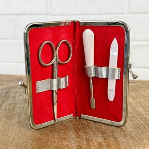 May include: Open silver-toned case with a red velvet interior. The case holds small scissors, a cuticle pusher, and a nail file. The tools are secured with silver straps. The case is open, revealing the tools inside.