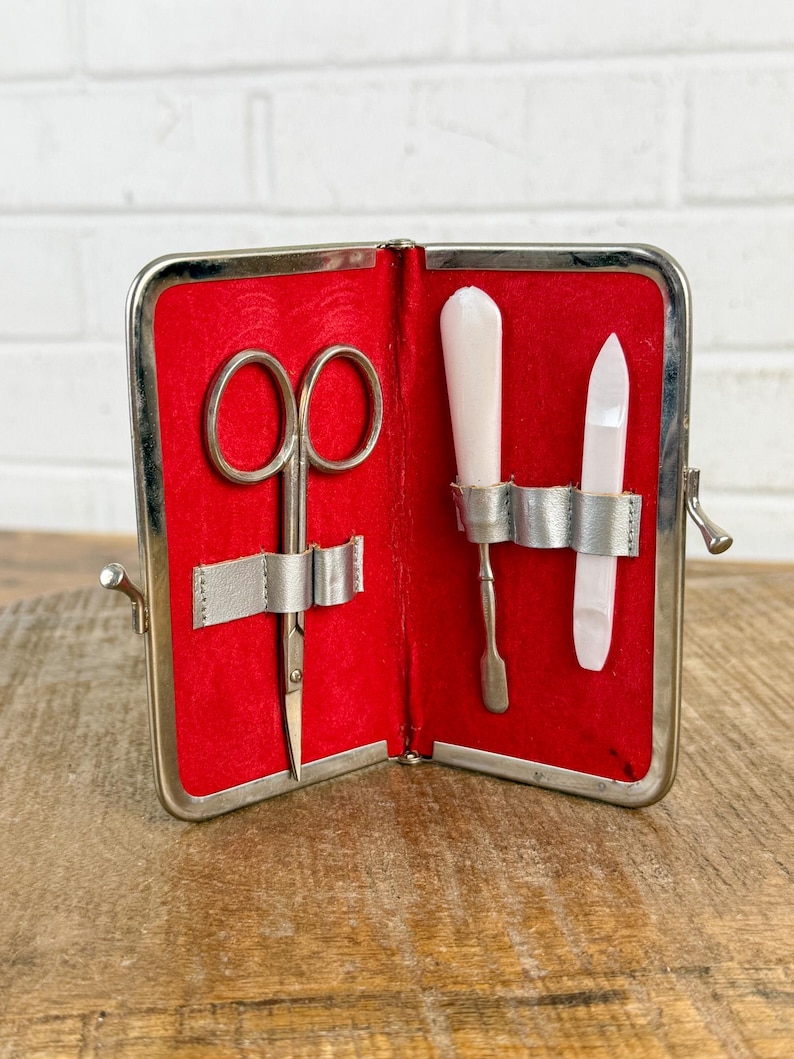 May include: Open silver-toned case with a red velvet interior. The case holds small scissors, a cuticle pusher, and a nail file. The tools are secured with silver straps. The case is open, revealing the tools inside.