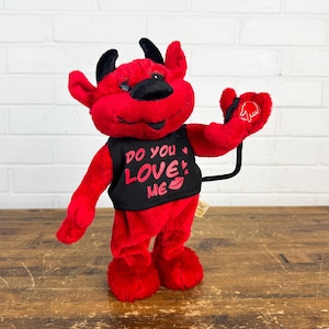 May include: A plush red devil figure with black horns, a black nose, and a black tail. It wears a black vest with the text "DO YOU LOVE ME" and a kiss mark. The devil has a heart-shaped hand gesture.