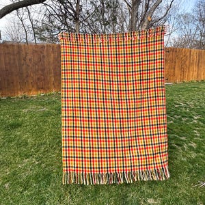 May include: A rectangular woven throw blanket featuring a repeating plaid pattern in vibrant shades of red, yellow, and dark navy blue. The blanket is finished with decorative fringe detailing along both the top and bottom horizontal edges.