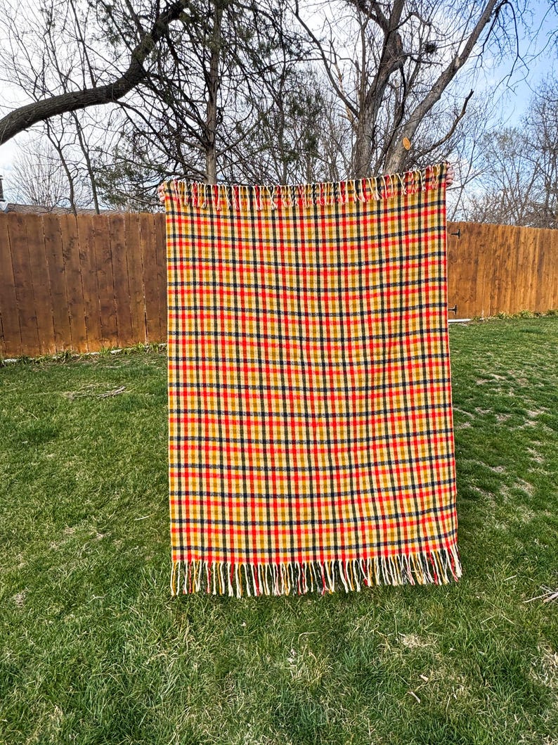 May include: A rectangular woven throw blanket featuring a repeating plaid pattern in vibrant shades of red, yellow, and dark navy blue. The blanket is finished with decorative fringe detailing along both the top and bottom horizontal edges.