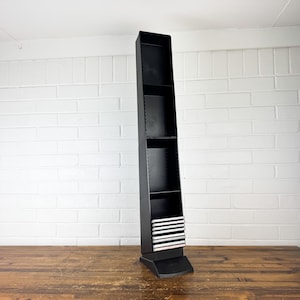 May include: A tall, black, freestanding shelf unit with multiple shelves. The unit is made of wood and has a rectangular shape. The shelf is placed on a wooden table against a white brick wall.
