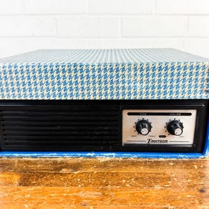 May include: Vintage Emerson radio with a black front panel and a blue and white houndstooth patterned top. The radio features tone and volume control knobs and the Emerson logo. The radio sits on a wooden surface.