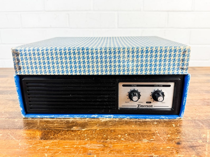 May include: Vintage Emerson radio with a black front panel and a blue and white houndstooth patterned top. The radio features tone and volume control knobs and the Emerson logo. The radio sits on a wooden surface.