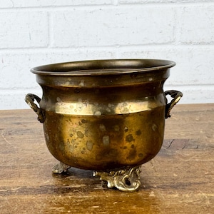 May include: A round brass pot with a weathered, tarnished finish. The vessel features two small, decorative side handles and rests on three ornate, curved feet. The metal surface shows visible signs of age, including dark spots and patina.