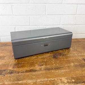 May include: A rectangular gray metal lockbox featuring a three-digit combination dial on the front panel. The box has a smooth, solid finish with a hinged lid and two small metal rivets visible near the top edge of the front closure mechanism.