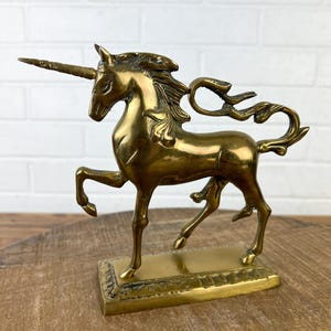 Puede incluir: Una figurita de unicornio de lat&oacute;n dorado con una melena y una cola flotantes. El unicornio est&aacute; de pie sobre una base rectangular con detalles decorativos.