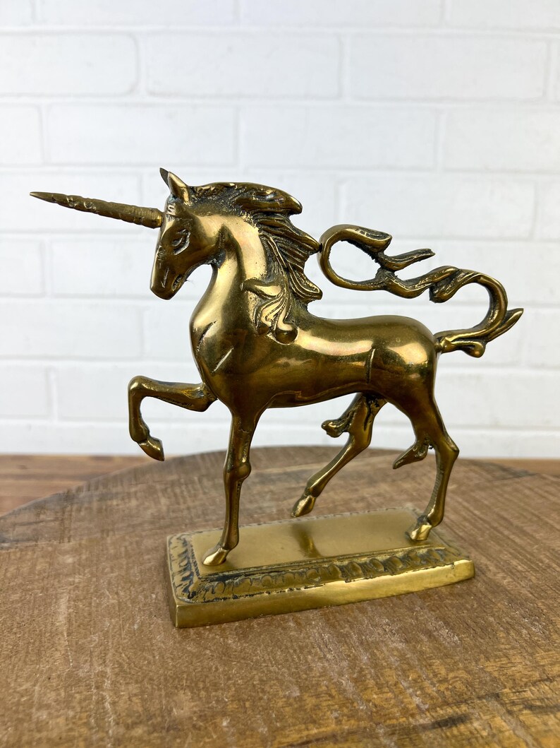 Puede incluir: Una figurita de unicornio de lat&oacute;n dorado con una melena y una cola flotantes. El unicornio est&aacute; de pie sobre una base rectangular con detalles decorativos.