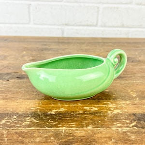 May include: A light green ceramic gravy boat with a handle. The boat has a curved shape and a spout for pouring. The handle is a loop design. The gravy boat is on a wooden surface.