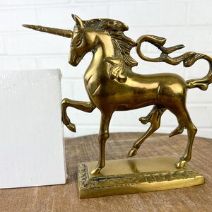 Puede incluir: Una figurita de unicornio de lat&oacute;n dorado con una melena y una cola fluidas. El unicornio est&aacute; de pie sobre una base rectangular con un borde decorativo.