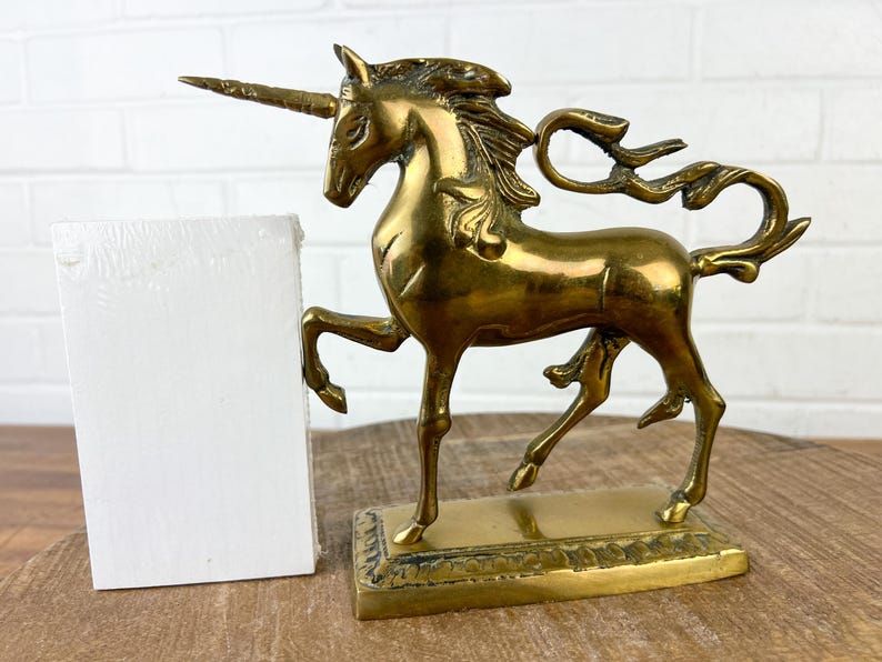 Puede incluir: Una figurita de unicornio de lat&oacute;n dorado con una melena y una cola fluidas. El unicornio est&aacute; de pie sobre una base rectangular con un borde decorativo.