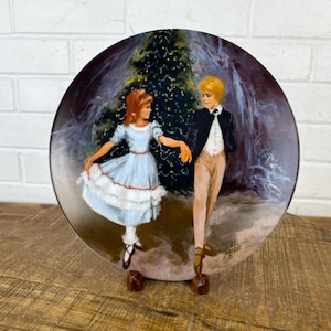 May include: A vintage Christmas plate featuring a couple dancing under a decorated Christmas tree. The woman is wearing a white dress with a red trim and the man is wearing a black suit with a white shirt. The plate has a dark background with a white and blue swirl design.