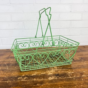 May include: A light green wire basket featuring a rectangular base and a repeating heart-shaped pattern along the sides. The basket includes a hinged, upright handle constructed from thin metal wire, providing a structured open-frame design.