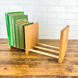 May include: A wooden book holder with a natural finish, holding a collection of green books. The holder has two vertical panels connected by three horizontal dowels. The books are various shades of green.
