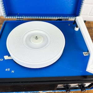 May include: A vintage blue portable record player with a white turntable and tonearm. The player has black knobs and a speaker grill. The open lid reveals a patterned interior. The player has settings for 33, 45, and N speeds.