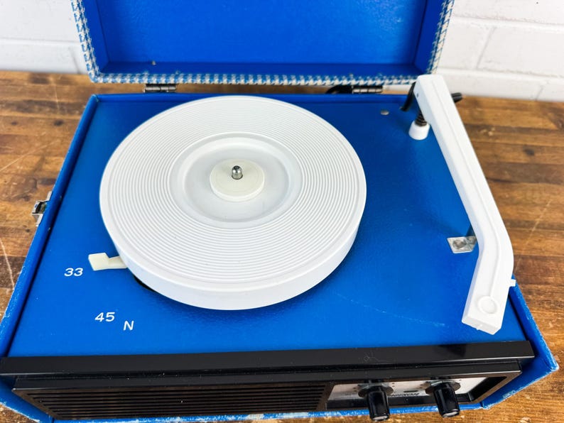 May include: A vintage blue portable record player with a white turntable and tonearm. The player has black knobs and a speaker grill. The open lid reveals a patterned interior. The player has settings for 33, 45, and N speeds.