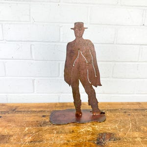 May include: A rustic metal sculpture of a person in a hat and poncho, standing on a small base. The sculpture is made of weathered, reddish-brown metal and is placed on a wooden surface against a white brick wall.