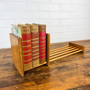 May include: Wooden book holder with three vintage books. The books have red and gold covers with text. The holder is made of light brown wood and has a built-in rack for additional books or display.