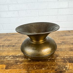 May include: A brass vessel with a rounded, bulbous body and a wide, flared rim. The metal surface displays a weathered, darkened patina with visible signs of age and oxidation, featuring a smooth, curved silhouette and a circular opening.