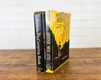 May include: Two vintage books on a wooden surface. The book on the left is titled "America's Camping Book" in white text on a black cover. The book on the right is titled "Survival With Style" with a yellow and black cover.