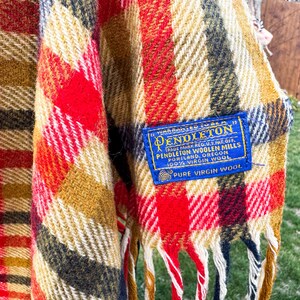 May include: A plaid blanket featuring a vibrant color palette of red, mustard yellow, black, and white. A blue rectangular fabric label is stitched onto the corner, reading: Warranted to be a Pendleton, Pendleton Woolen Mills, 100% Virgin Wool.
