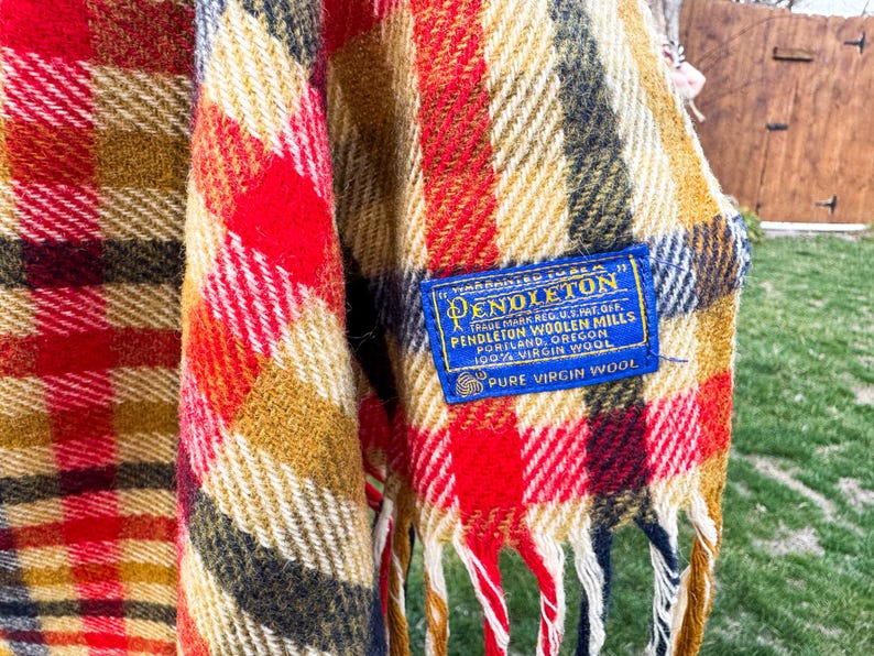 May include: A plaid blanket featuring a vibrant color palette of red, mustard yellow, black, and white. A blue rectangular fabric label is stitched onto the corner, reading: Warranted to be a Pendleton, Pendleton Woolen Mills, 100% Virgin Wool.