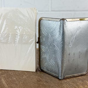 May include: A vintage silver cigarette case with an ornate floral design and a stack of white cards wrapped in clear plastic. The case has a hinged opening and a clasp. The items are on a wooden surface.