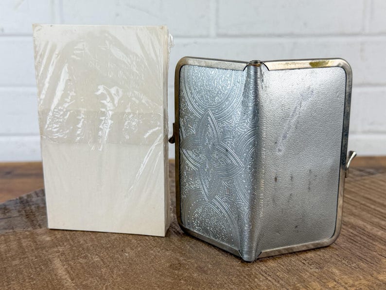 May include: A vintage silver cigarette case with an ornate floral design and a stack of white cards wrapped in clear plastic. The case has a hinged opening and a clasp. The items are on a wooden surface.