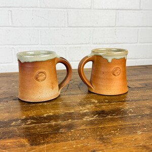 May include: Two handmade ceramic mugs with a rustic, earthy aesthetic. The mugs are a warm brown color with a lighter, off-white rim. Each mug features a small, embossed design and a curved handle. The mugs are displayed on a wooden surface.