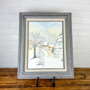 May include: A framed watercolor painting of a winter landscape. The artwork depicts a snow-covered scene with a house, trees, and a path. The frame is gray, and the painting is displayed on a wooden surface.