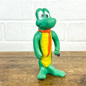 May include: A green alligator figurine with a yellow belly and orange tie. The alligator has white eyes and a black object in its right hand. The figurine is standing on a wooden surface.