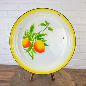 Puede incluir: Una bandeja de servir de esmalte blanco con un borde amarillo y un dise&ntilde;o pintado de naranjas en una rama. La bandeja est&aacute; sobre un soporte de madera.